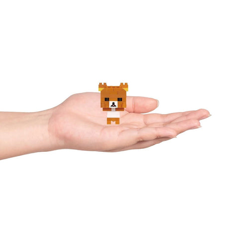 A hand holds a small, blocky toy bear from the Nanoblock Rilakkuma Blind Bag - Vol. 3 series, featuring a brown head and body, black eyes and nose, and a white belly. Its pixelated design against the plain white background entices you to collect all six in this collection by Nanoblock.