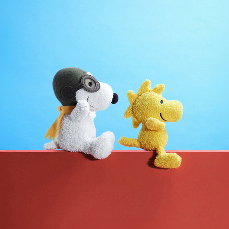 The Snoopy Plush Flying Ace, an 18cm white plush toy from the Snoopy brand featuring goggles, is seated alongside a yellow bird on a red surface against a blue background, capturing the delightful essence of Peanuts Gift.