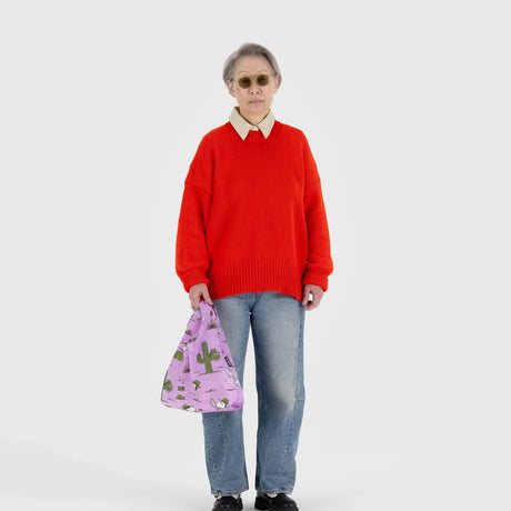 A person wearing a red sweater, jeans, and sunglasses stands against a plain white background, holding the Baggu Baby Baggu Peanuts - Snoopy Pink Spike bag featuring cactus designs and crafted from recycled nylon.