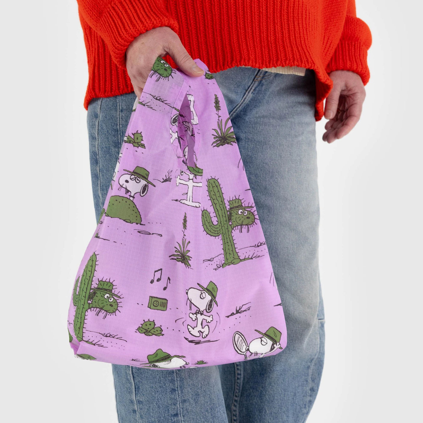 A person in an orange sweater and jeans holds the Baby Baggu Peanuts - Snoopy Pink Spike by Baggu, a purple bag made from recycled nylon, featuring playful cartoon cactus and dog illustrations.