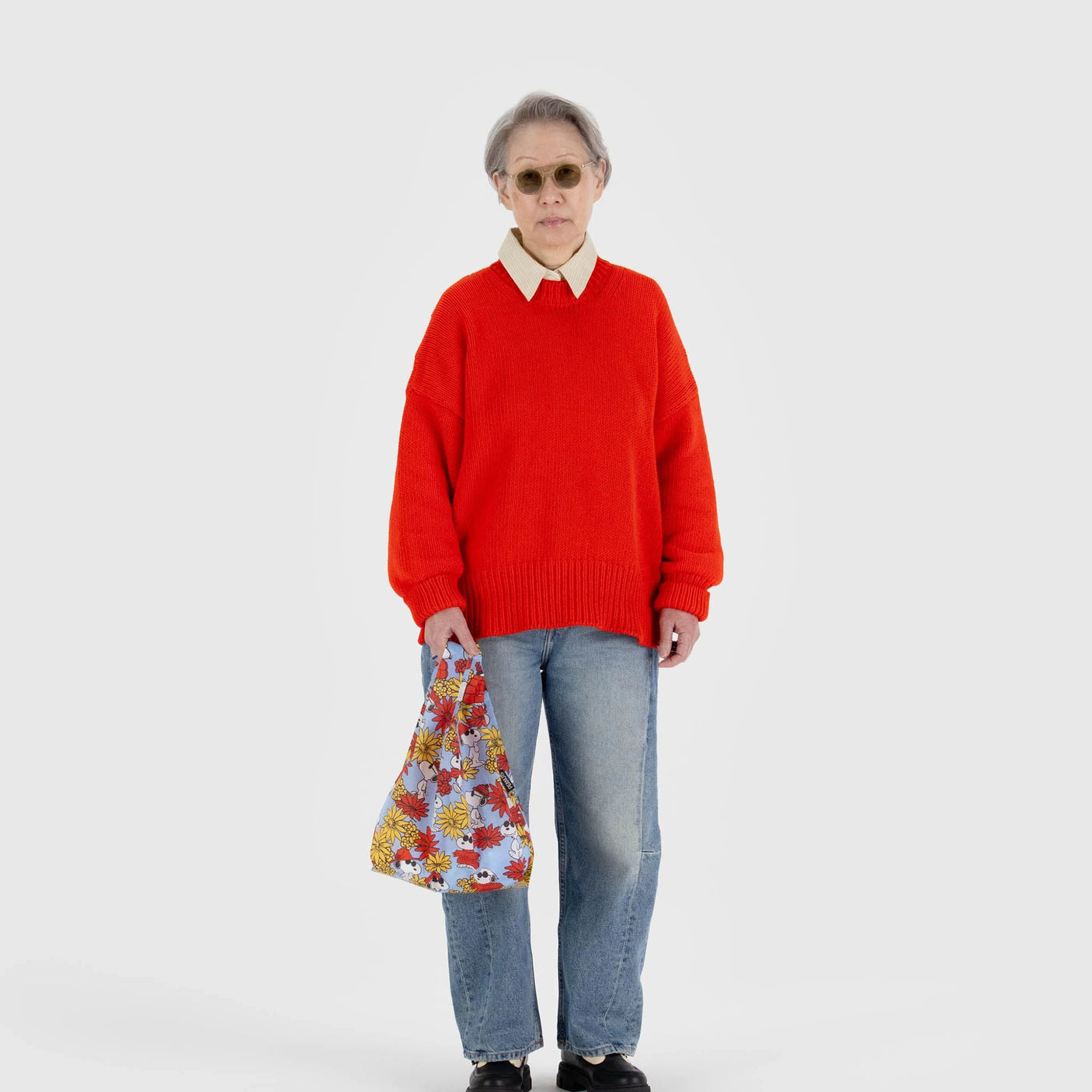 A person in a red sweater, jeans, sunglasses, and black shoes stands against a plain white background, holding a Baby Baggu Peanuts - Floral Snoopy from Baggu, crafted from recycled nylon.