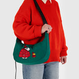 A person in an orange sweater and jeans is holding the Baggu x Peanuts Medium Crescent Bag, featuring embroidered Snoopy.