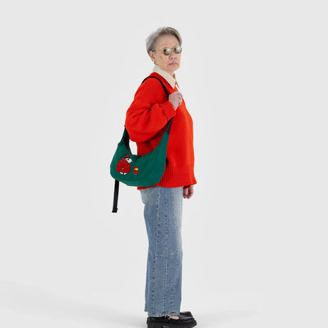 A person in an orange sweater and jeans stands sideways, holding a Baggu x Peanuts - Medium Crescent Bag Embroidered Snoopy.