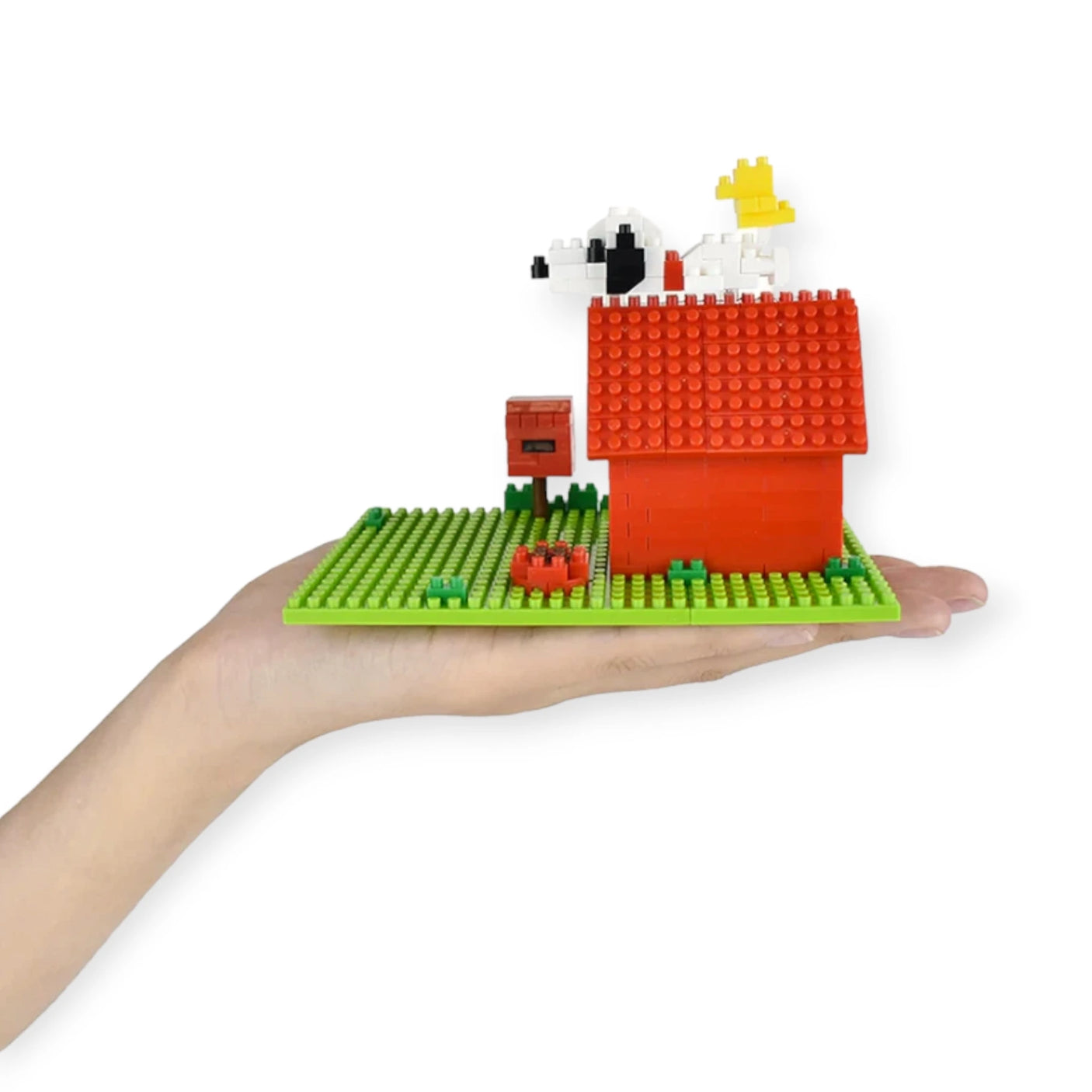 A hand presents a finely crafted Nanoblock Peanuts - Snoopy House Set, showcasing a red house complete with a mailbox and Snoopy relaxing on the roof. Against the plain white background, this delightful design honors Peanuts history with its micro-sized blocks characteristic of the renowned Nanoblock brand.