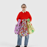 A person wearing a red sweater and jeans holds a set of three vibrant and patterned Peanuts x Baggu Standard Bags, crafted from recycled nylon, against a plain background.