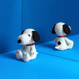 A Snoopy Plush Sitting 19cm - Corduroy Cream, featuring black ears and a red collar, rests on a bright blue surface, facing a mirrored wall. Perfect for any Peanuts enthusiast, this delightful Snoopy stuffed dog toy embodies whimsical joy and nostalgia.