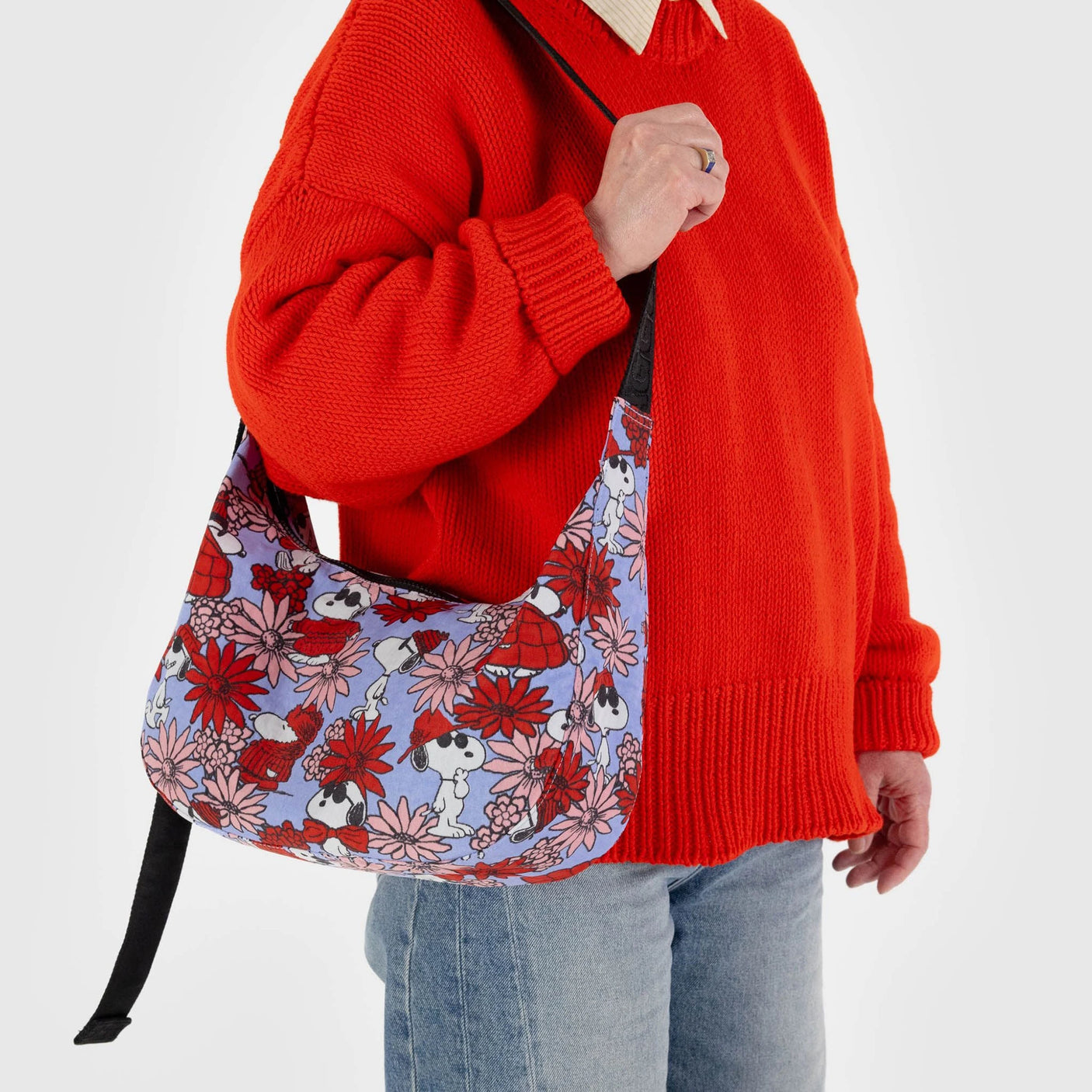 A model in a red sweater displays the Baggu x Peanuts Medium Crescent Bag, featuring a vivid floral Snoopy design and made from recycled nylon.