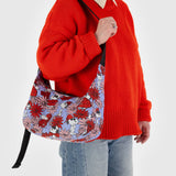 A model in a red sweater displays the Baggu x Peanuts Medium Crescent Bag, featuring a vivid floral Snoopy design and made from recycled nylon.