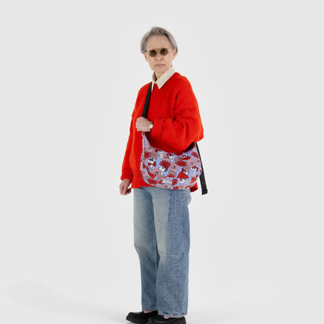 A person dressed in a red sweater, jeans, and sunglasses is holding the Baggu x Peanuts - Medium Crescent Bag Floral Snoopy, a vibrant recycled nylon shoulder bag featuring a playful Snoopy design.