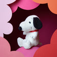 A Snoopy Plush Sitting 20cm - Terry Cream toy dog, featuring a red collar, is set against a pink and red cloud-patterned background, showcasing its soft terry cloth exterior and eco-friendly recycled polyester filling.