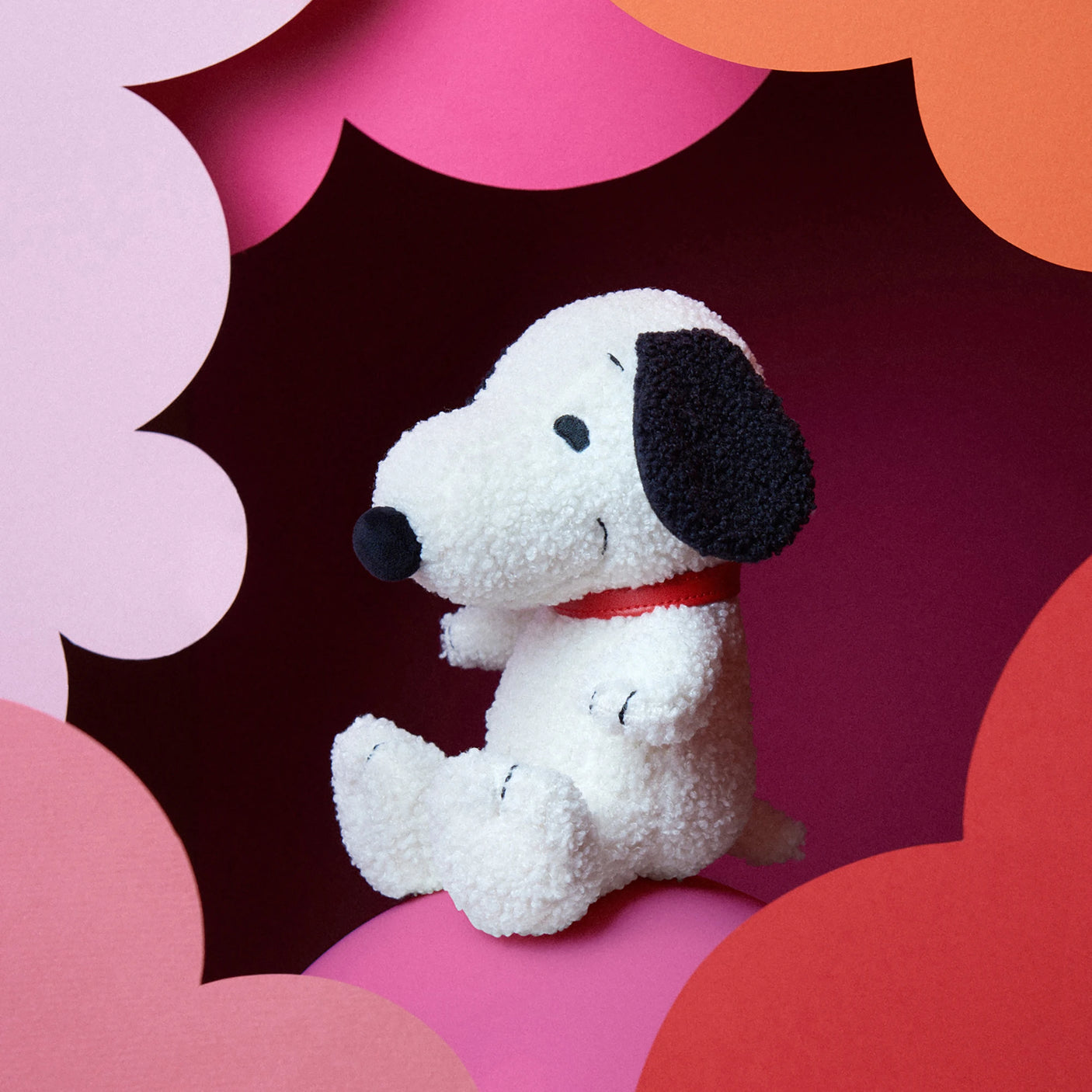 A Snoopy Plush Sitting 20cm - Terry Cream toy dog, featuring a red collar, is set against a pink and red cloud-patterned background, showcasing its soft terry cloth exterior and eco-friendly recycled polyester filling.