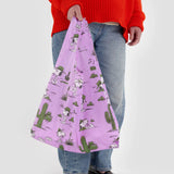 A person carries the Baggu Standard - Peanuts Pink Spike, an eco-friendly bag featuring vibrant cactus and cartoon dog designs. They are wearing a red sweater and jeans.