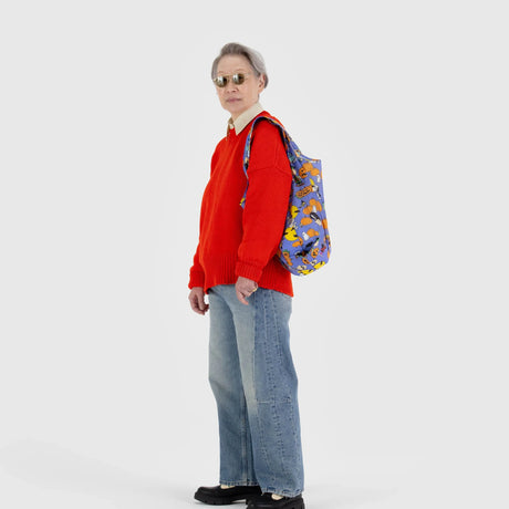 Person wearing a red sweater, blue jeans, and sunglasses faces left, carrying a Baggu Standard Baggu x Peanuts - Pumpkin Patch backpack over one shoulder against a plain white background.
