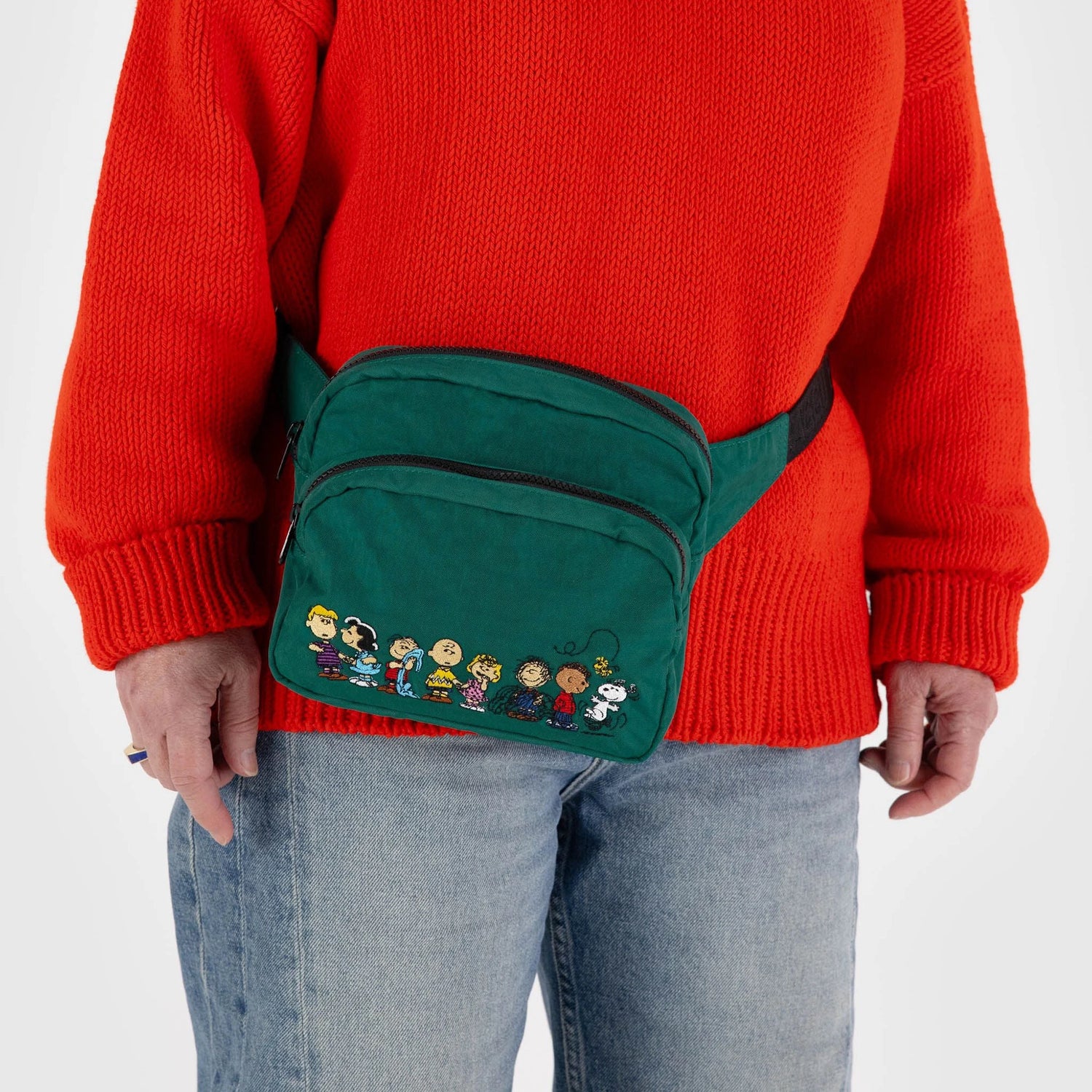 A person wearing a bright orange sweater, light blue jeans, and a green Peanuts x Baggu Fanny Pack adorned with cartoon characters. This limited edition accessory is made from recycled nylon, contributing an eco-friendly element to their colorful outfit.