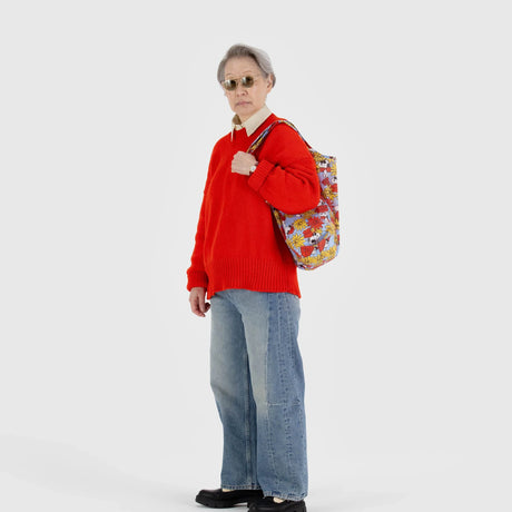 An older adult wearing sunglasses, a red sweater, and blue jeans stands against a plain background, with the Baggu Standard - Peanuts Floral Snoopy tote bag crafted from recycled nylon draped over their shoulder.