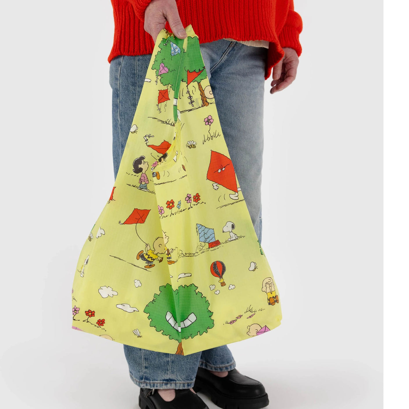 A person carries the Baggu Standard - Peanuts Kite Eating Tree tote bag, showcasing cartoon characters and kites, while wearing a red sweater and jeans.