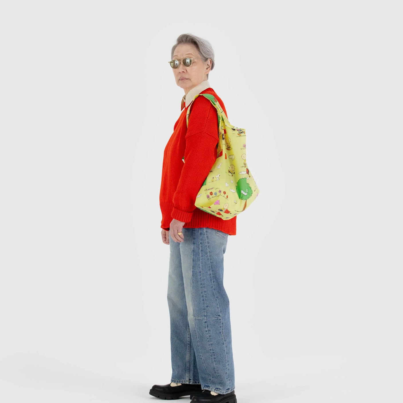 Against a plain white background, a person wearing sunglasses, a red sweater, blue jeans, and black shoes carries a Baggu Standard - Peanuts Kite Eating Tree tote bag featuring a yellow pattern on their shoulder.