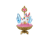 A figure from the Re-Ment Pokemon Gemstone Collection 2, featuring a creature with pink and white accents, is displayed in a gold bowl filled with pink crystals. The scene includes blue crystal-like structures and a gold staff topped with a pink gem.