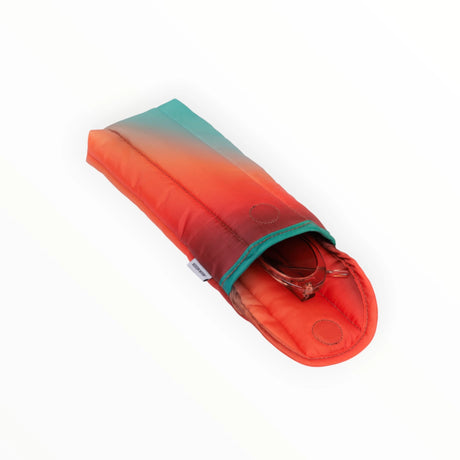 The Julia Heuer x Baggu Puffy Glasses Sleeve - Rain, crafted by Baggu, transitions from orange to teal in a vibrant gradient. Made from recycled materials, the partially open sleeve reveals glasses inside against a simple white background.