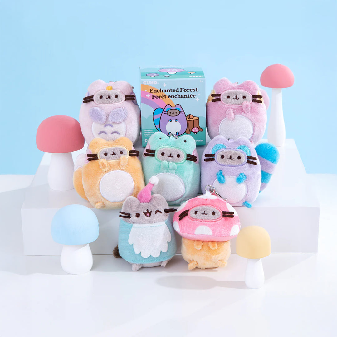 A set of eight Pusheen Blind Box - Enchanted Forest plush toys by Pusheen is displayed in two rows on a white platform, surrounded by pastel mushroom props, a small box, and a cute plush keychain in the background.