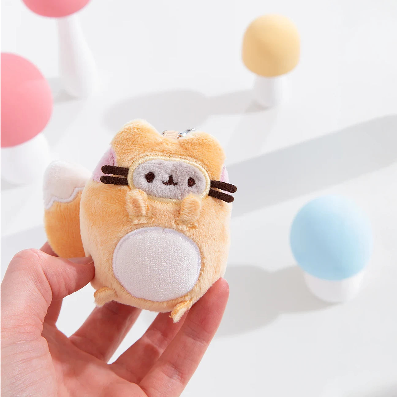 A hand holds a small, round, orange and white Pusheen plush toy with a mask-like face from the Pusheen Blind Box - Enchanted Forest collection. Colorful blurred objects in the background hint at more plush keychains in the series.