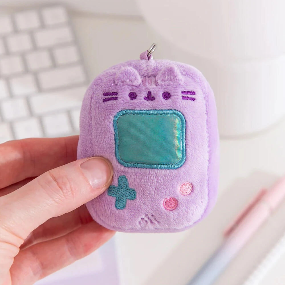 A hand holds a Pusheen Blind Box - Gamer Series keychain by Pusheen, shaped like a retro handheld game console with embroidered cat features in soft purple plush.