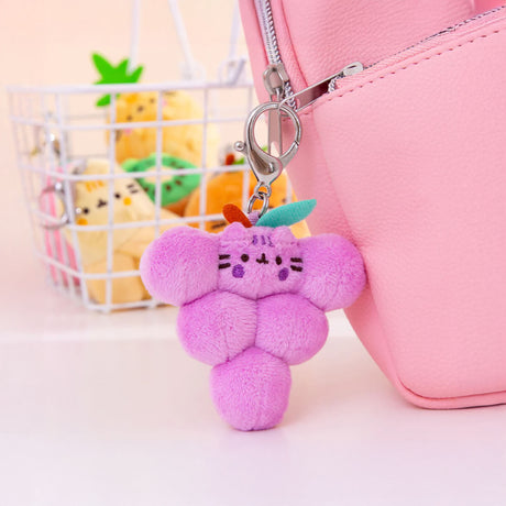 A pink backpack is adorned with a plush grape keychain, a delightful fruity plush that adds charm. In the background, there's a white basket holding other plush fruit toys from the Pusheen Blind Box - Fruits Series by Pusheen, creating a scene reminiscent of delightful treasures.