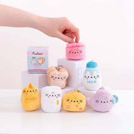 A hand arranges mini plushies from the Pusheen Blind Box - Kitchen Surprise Series by Pusheen, featuring designs like milk, honey, and a teapot. These charming figures are creatively displayed on white blocks against a simple background.