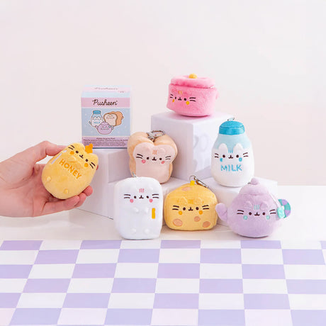 A hand holds a plush toy labeled "Honey" amidst six small, pastel-colored mini plushies from the Pusheen Blind Box - Kitchen Surprise Series by Pusheen, displayed on square platforms atop a checkered surface.