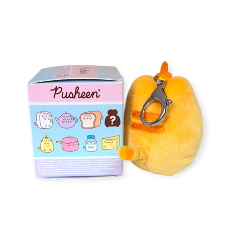 This plush yellow keychain with an orange stripe makes an ideal addition to the Pusheen Blind Box - Kitchen Surprise Series. It's displayed beside a Pusheen box adorned with illustrations of various cat-themed items.