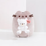 This adorable plush toy, inspired by Pusheen, is shaped like a gray cat and holds a small white GUND bear. It features a unique pink "GUND" tag and is named the Pusheen x GUND Kai Bear Plush - 24cm - Limited Edition.