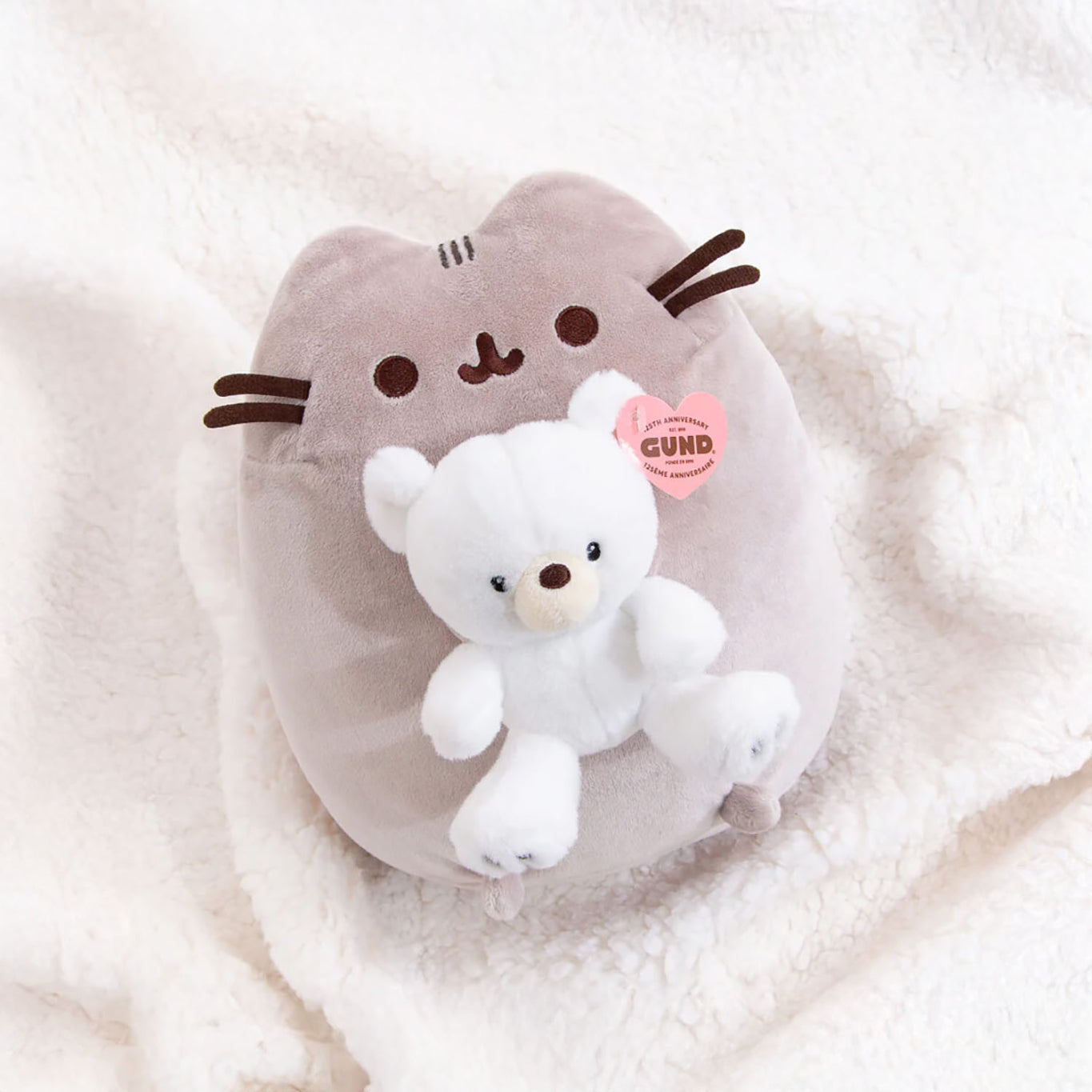 A plush cat pillow with whiskers, similar to a Pusheen Plush, holds a charming new companion—the limited edition Pusheen x GUND Kai Bear Plush—both nestled on a soft, cream-colored blanket.