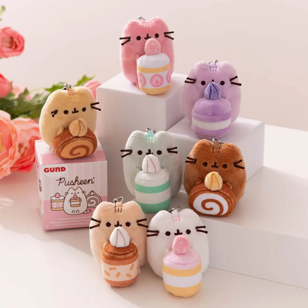 Six small Pusheen plush cats from the Sweets Series Blind Box hold pastel treats on white blocks, with a pink box and flowers nearby—ideal for collectors of Pusheen or anyone who loves cute dessert-themed keychains.