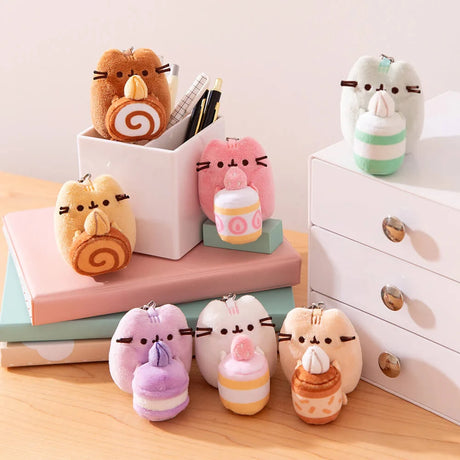 Seven adorable Pusheen Sweets Series plush keychains by Pusheen, each holding a dessert-themed treat, are displayed on stacked books and a white drawer unit atop a wooden surface.
