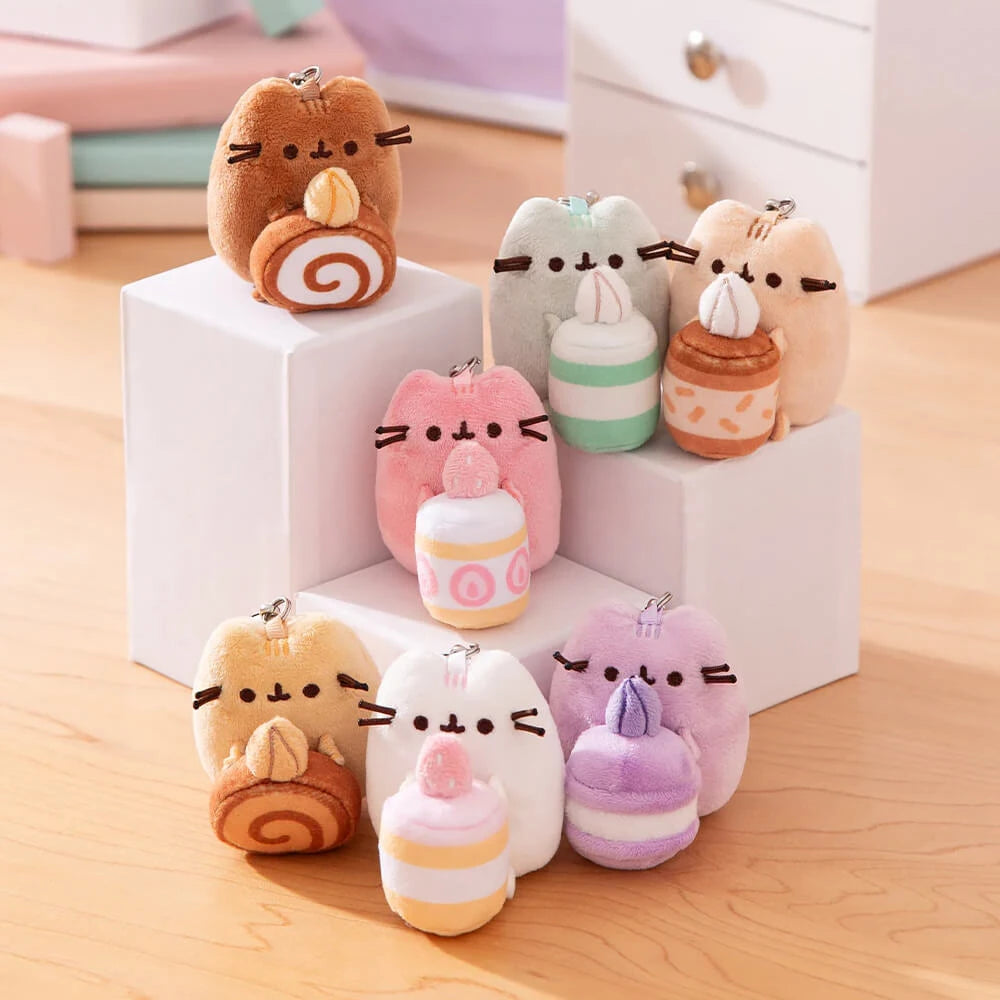 Nine pastel Pusheen Blind Box - Sweets Series plush keychains by Pusheen, each featuring a cute cat and dessert, are displayed on white blocks and a wooden surface.
