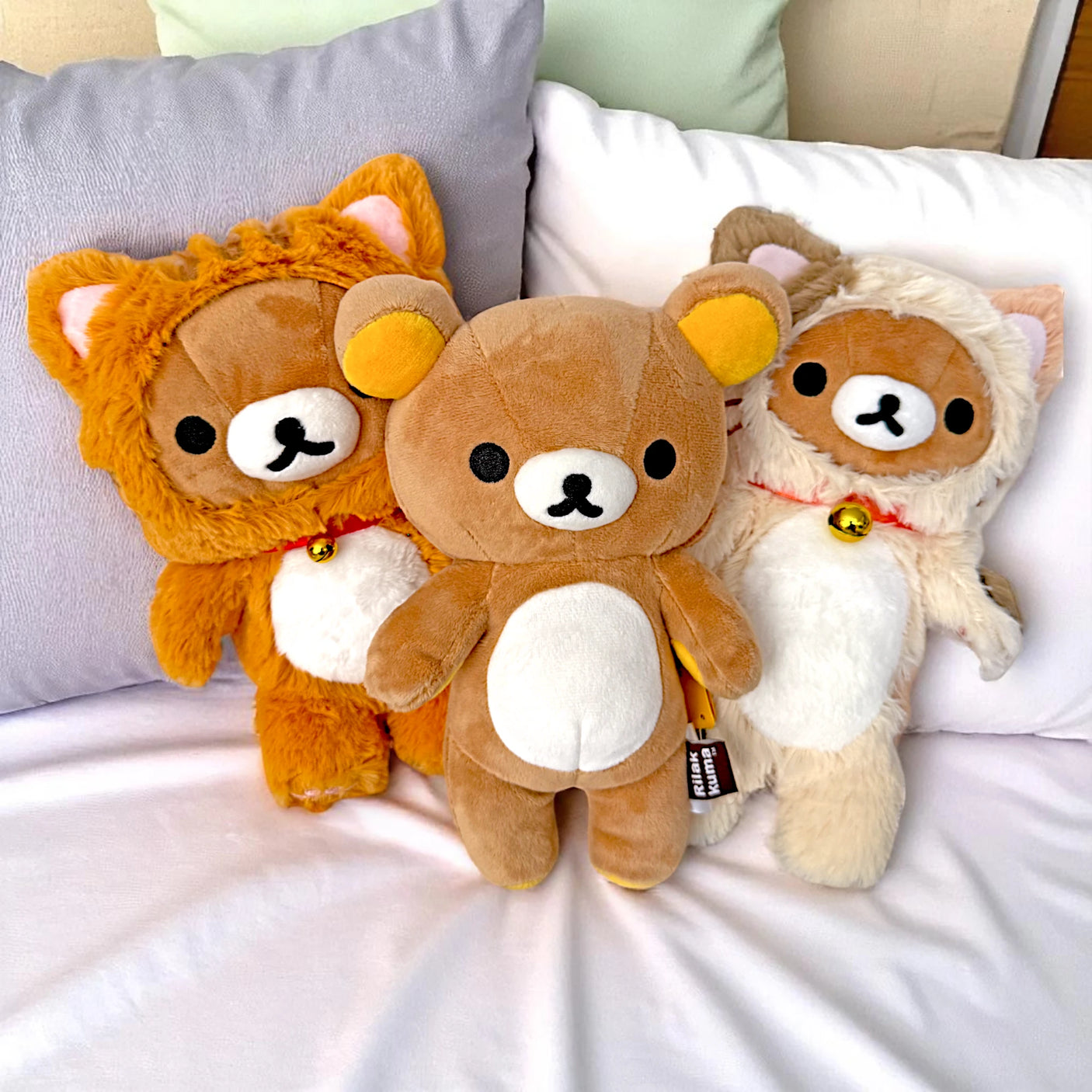 Three plush teddy bears sit on a bed with white sheets and pillows; two wear animal costumes, while the Rilakkuma Small Plush by Rilakkuma features a hidden zipper pouch for storing tiny treasures.