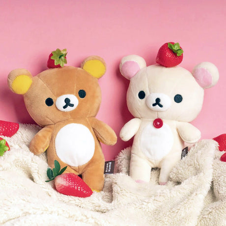 Two Rilakkuma Small Plush bears, one brown and one cream, each feature a strawberry on their head and a hidden zipper pouch. They sit on a soft beige blanket with scattered strawberries against a pink background. Brand: Rilakkuma.