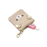 A small Rilakkuma Coin Pouch Keychain from the Rilakkuma brand, featuring a beige bear face design, is slightly unzipped to show a pink tube of hand sanitizer. A gold clip hangs from the synthetic leather pouch, adding charm.