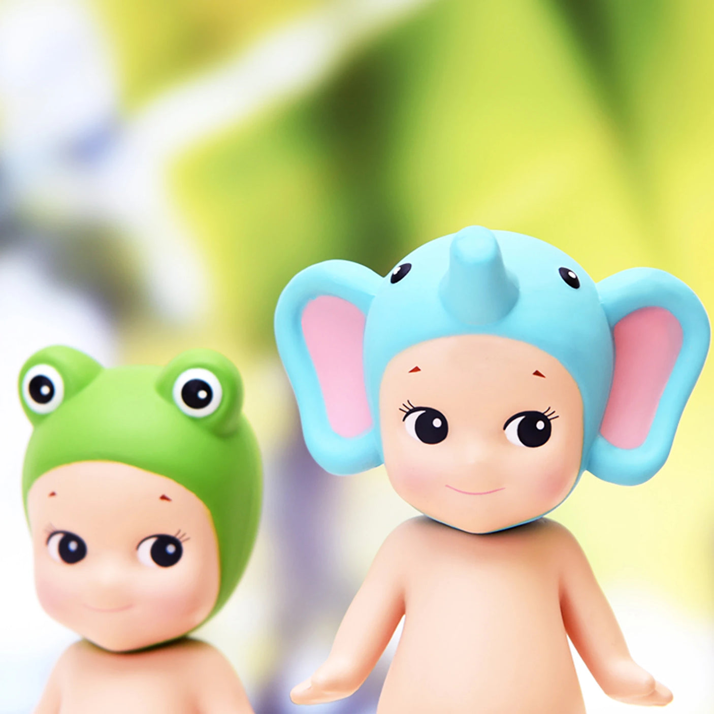 Two adorable dolls from the Sonny Angel Animal Version 1 Series by Sonny Angel, one sporting a green frog hat and the other a blue elephant hat, stand charmingly against a softly blurred green backdrop. Ideal collectibles for fans of delightful miniatures.