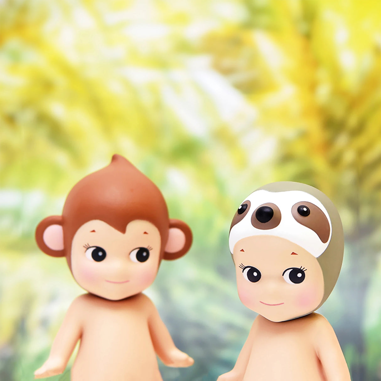 Two toy figures from the Sonny Angel Animal Version 1 Series by Sonny Angel, each donning whimsical animal-themed hats—one portraying a lively monkey and the other depicting a tranquil sloth—pose against a blurred spectrum of colors, showcasing the charm of these enchanting collectibles.