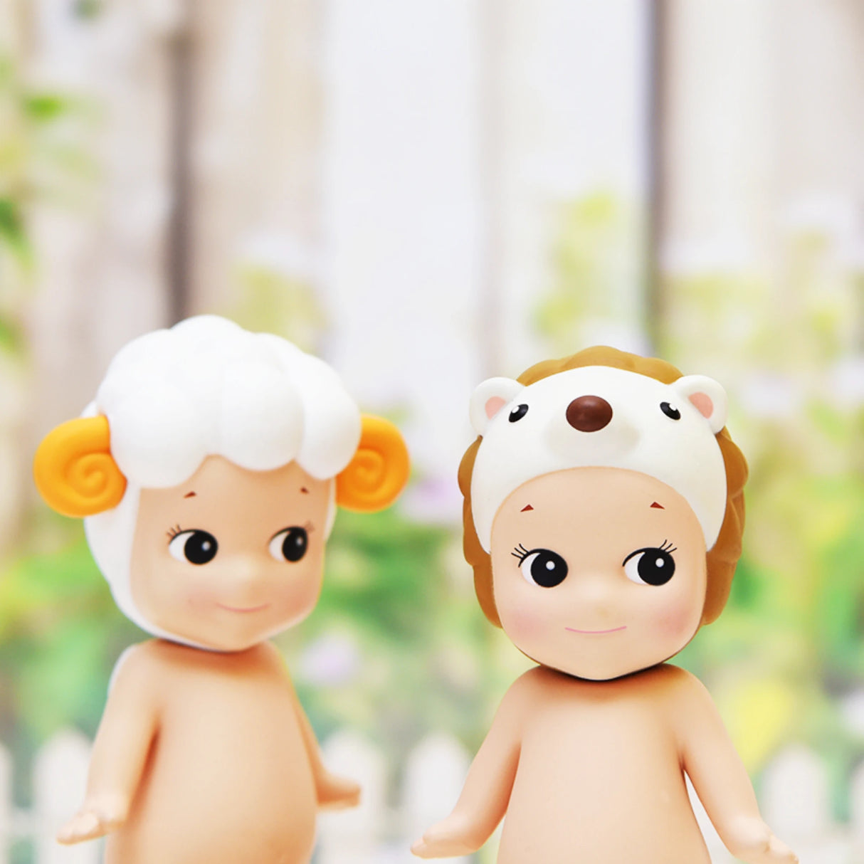 Two Sonny Angel figurines from the Sonny Angel Animal Version 2 Series stand side by side, each emerging from a blind box. One is adorned with a sheep hat featuring yellow horns, and the other wears a hedgehog hat. Both display cheerful smiles against a softly blurred background.