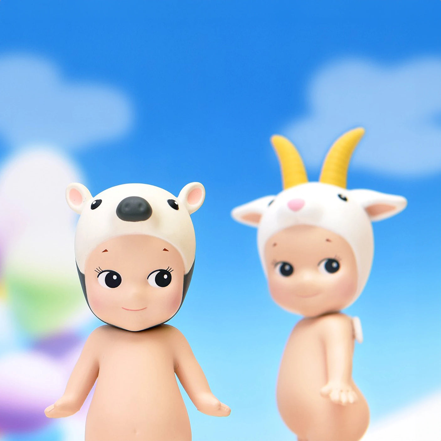 Two Sonny Angel Animal Version 4 Series dolls, one sporting a white bear hat and the other wearing a goat hat, stand against a backdrop of a blue sky with clouds, beautifully showcasing the charm of these collectible figures from Sonny Angel.