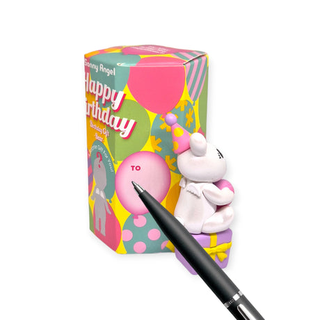 A petite Sonny Angel Birthday Bear, dressed in a unicorn outfit, is affixed to a vibrant gift box adorned with "Happy Birthday" text, with a black pen pointing at the box.