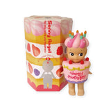 A Sonny Angel Happy Birthday Cake Gift figurine, adorned with a festive birthday cake hat complete with candles and holding a "Happy Birthday" cake, stands happily beside its colorful blind box.