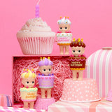 Four figurines from the Sonny Angel Happy Birthday Cake Gift series, each with a "Happy Birthday" sign, are placed on and around pink and white gift boxes. A large decorative cupcake and a Sonny Angel figure stand proudly in the background.