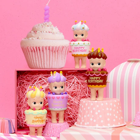 Four figurines from the Sonny Angel Happy Birthday Cake Gift series, each with a "Happy Birthday" sign, are placed on and around pink and white gift boxes. A large decorative cupcake and a Sonny Angel figure stand proudly in the background.