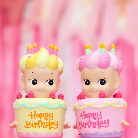 Two Sonny Angel Happy Birthday Cake Gift dolls, from the Sonny Angel brand, wear cupcake hats decorated with candles and frosting. Standing together, they each sport a pastel outfit inscribed with "Happy Birthday," making them a charming surprise for any fan of blind box collectibles.