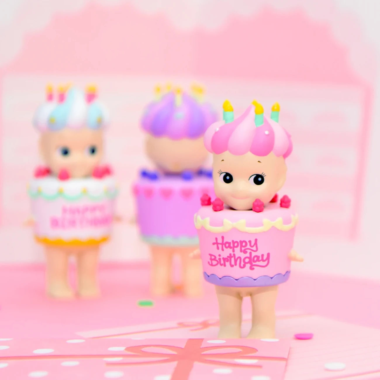 A set of three Sonny Angel Happy Birthday Cake Gift figures from the Sonny Angel brand, each styled as a cupcake with "Happy Birthday" inscribed on them, displayed against a pink background.