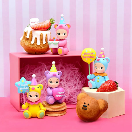 The Sonny Angel Birthday Bear Gift figurines by Sonny Angel are adorned in colorful outfits and hold cheerful signs, complemented by a backdrop of pink decorations, pastries, and strawberries on a striped setting. These delightful gifts bring an extra burst of joy to any celebration.