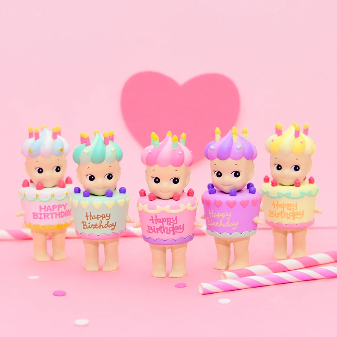 Five Sonny Angel Happy Birthday Cake Gift figurines, adorned with vibrant hats, are lined up against a pink backdrop featuring a heart shape. Two striped straws rest in front, inviting you to embrace the whimsical charm of the Sonny Angel brand's Birthday Cake Series.